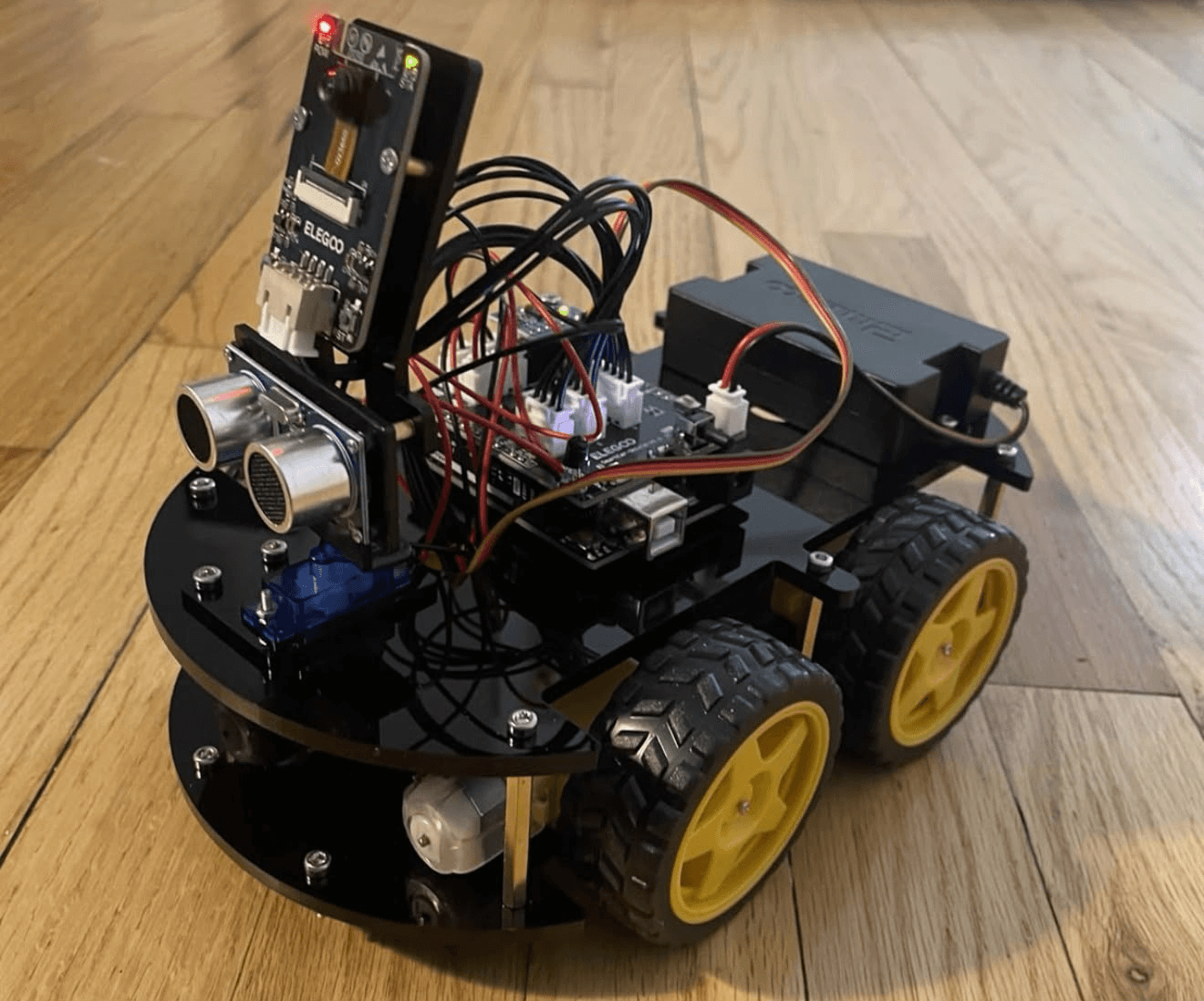 RC-X four-wheel robot platform on wood floor: black acrylic chassis, yellow wheels, ELEGOO control board, ultrasonic sensor on pan servo, camera module
